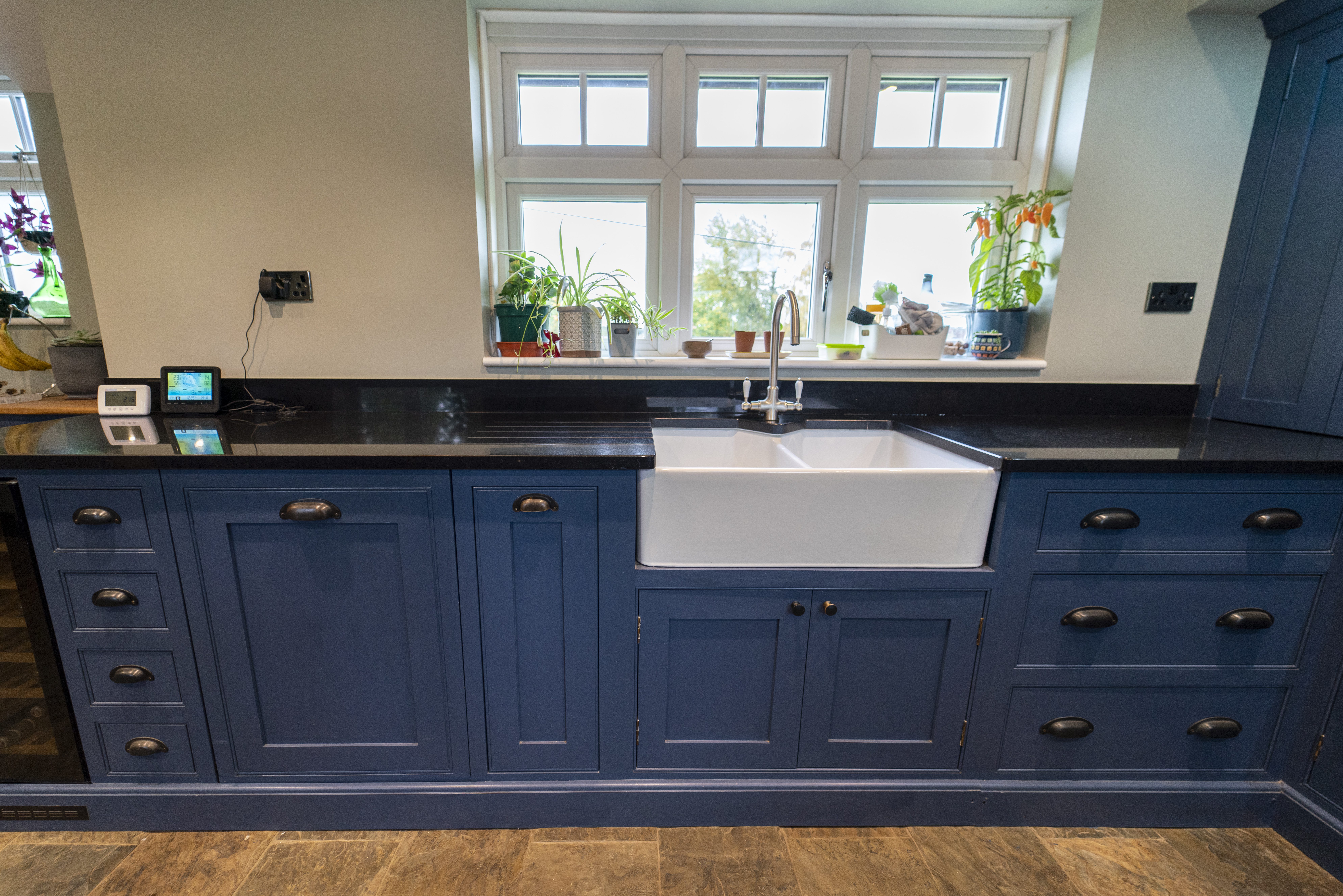 Front view of a blue kitchen sink run with Absolute Black Granite worktops, showing a white farmhouse sink, tap and window plants above the worktop.