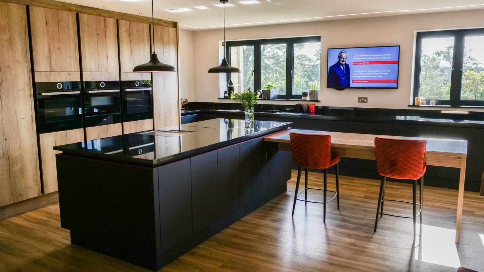 Wide angle view of a contemporary kitchen with an Absolute Black Granite island worktop and breakfast bar, showing bar stools, timber cabinetry and large windows.